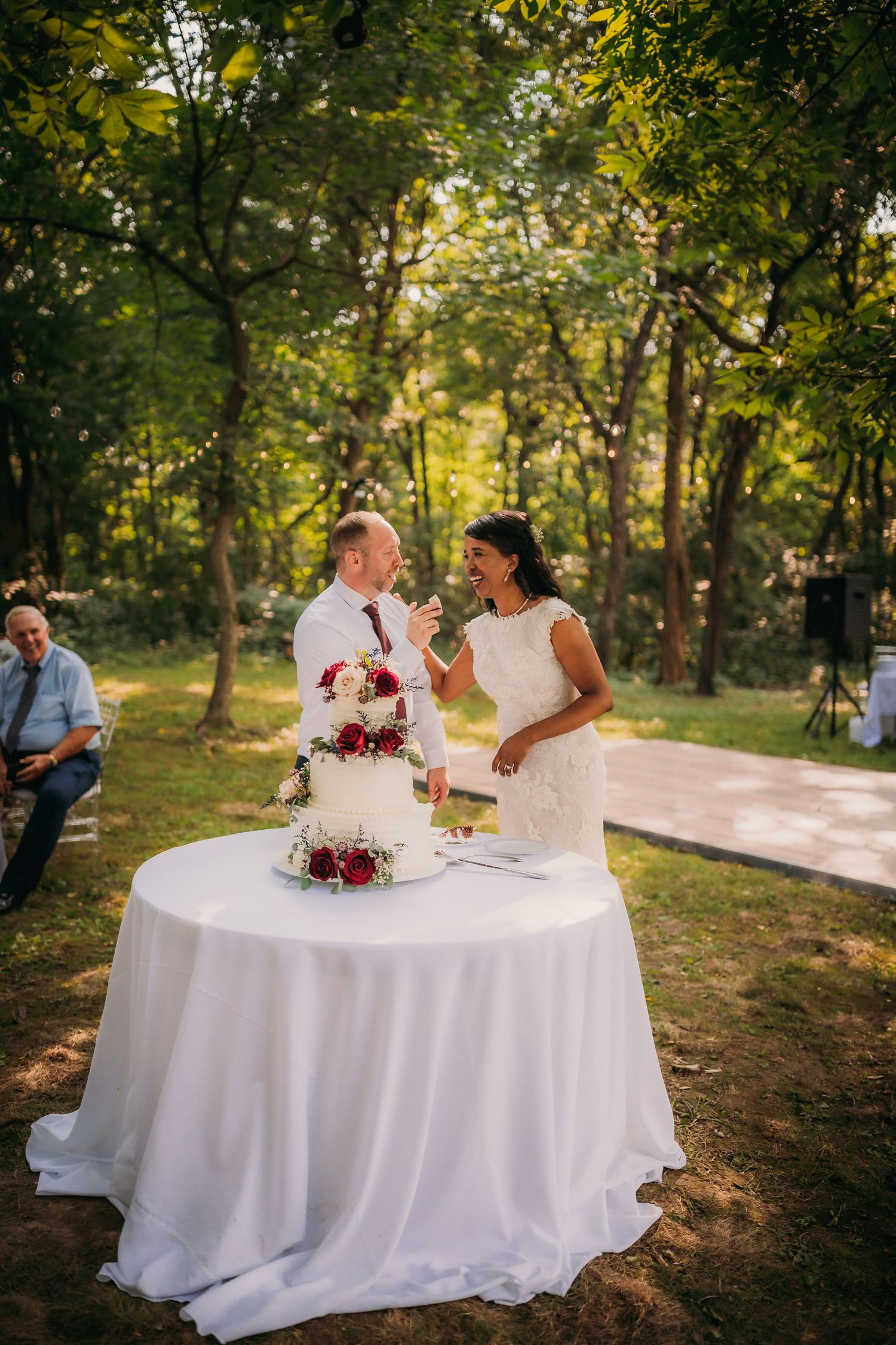 Newlyweds share a moment while cutting their elegant white and burgundy wedding cake at an outdoor forest reception.