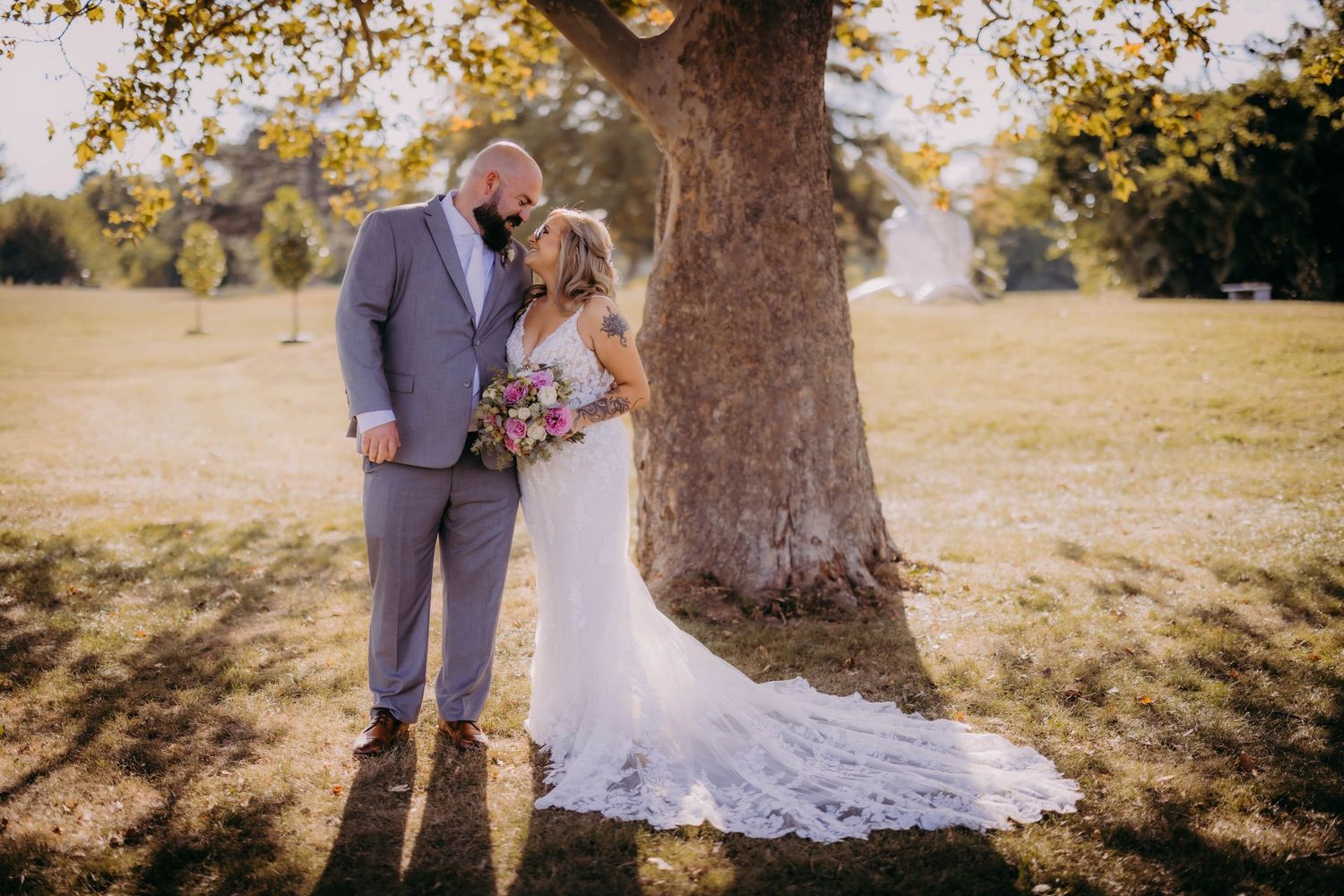 A romantic wedding photo of a couple embracing under a large tree with sunlit golden foliage.