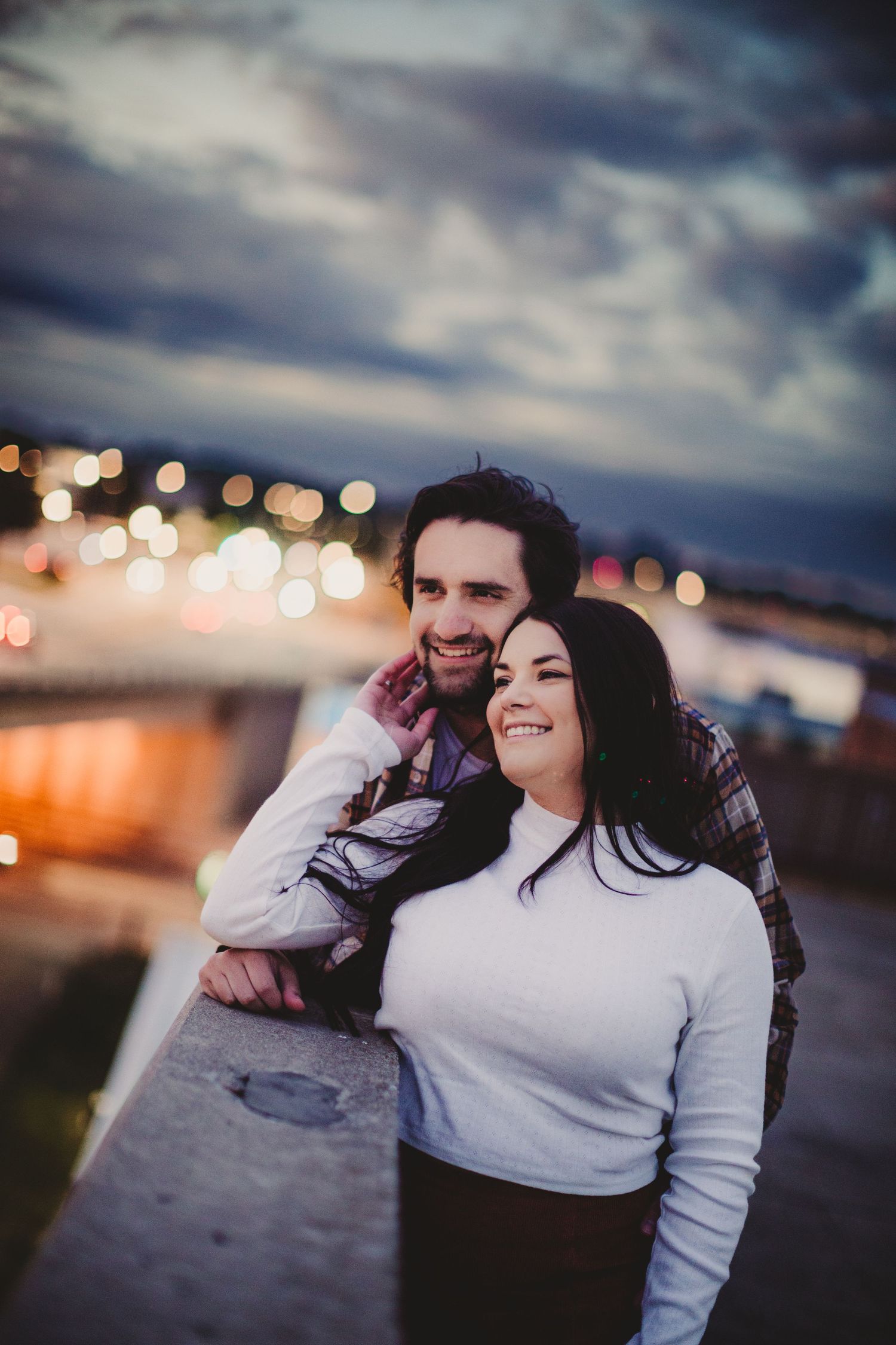 A couple smiles while leaning against a railing with bokeh city lights in the background.