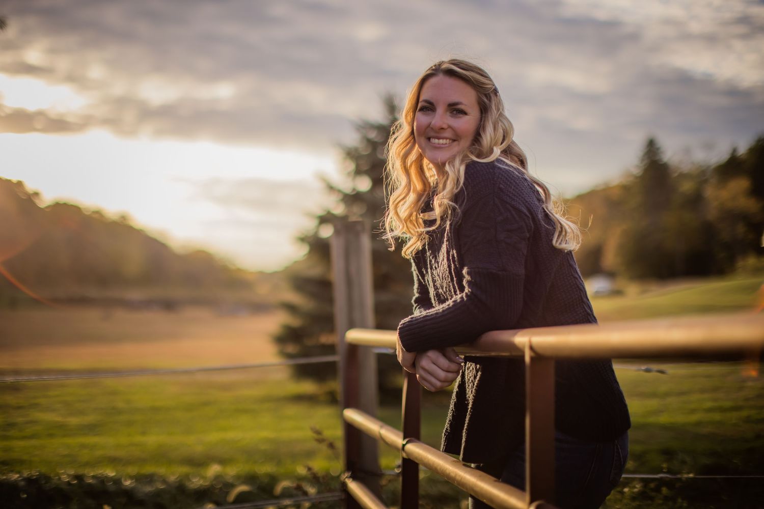 Woman in dark sweater smiles while leaning on wooden fence at sunset.