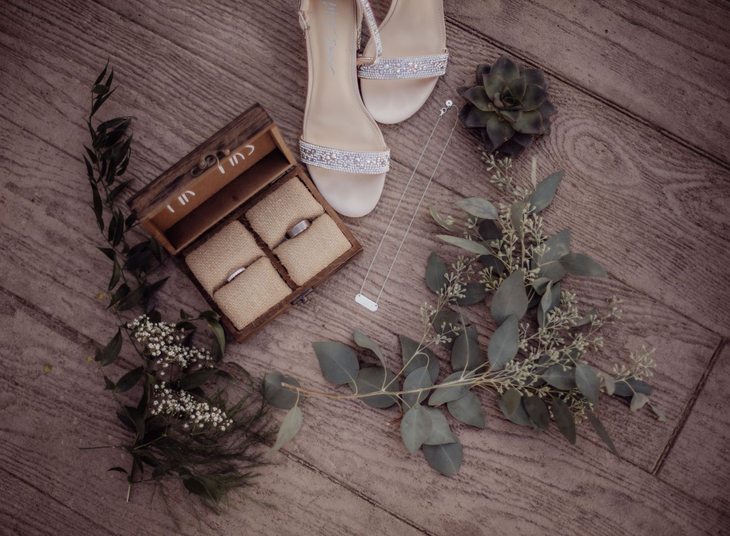 Overhead view of wedding shoes, ring box and eucalyptus branches on wood flooring.