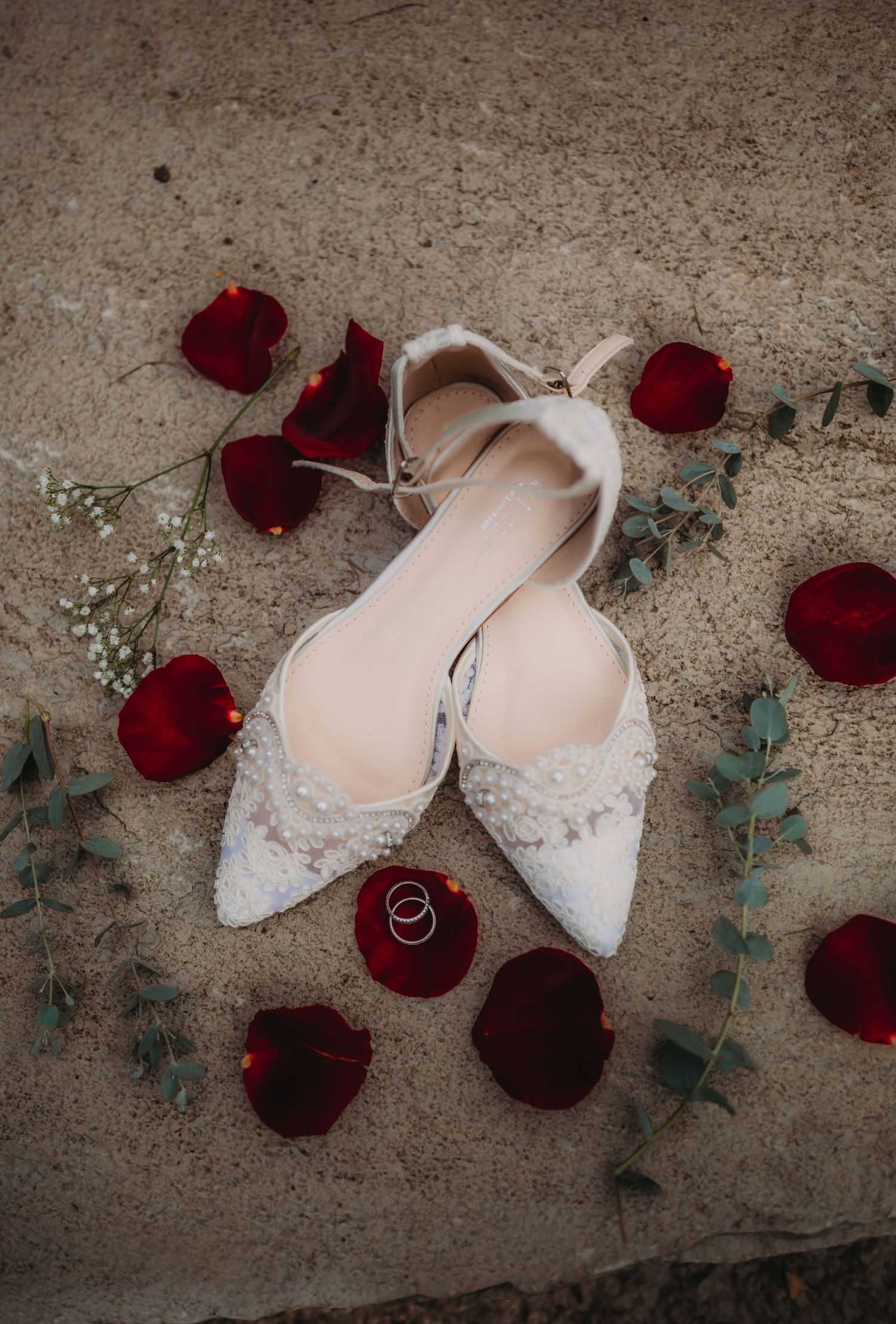 Elegant lace bridal flats surrounded by deep red rose petals and eucalyptus leaves on concrete background.