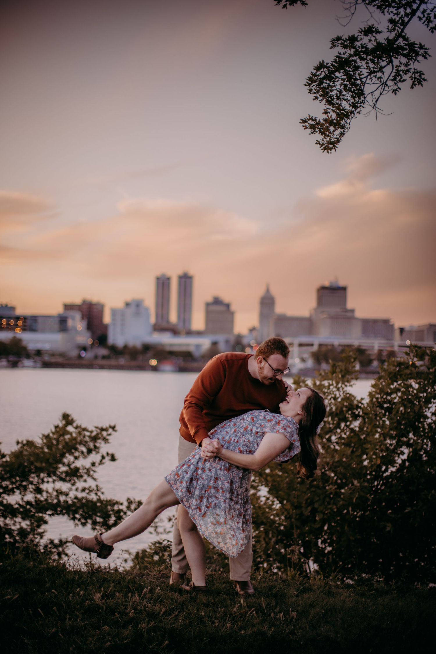Couple performs romantic dance dip pose with city buildings and sunset sky in background.