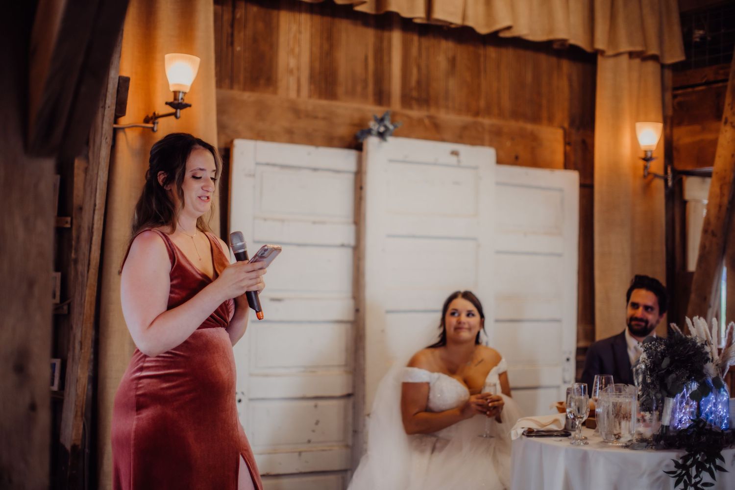Wedding reception features speaker in burgundy dress addressing seated guests in rustic barn venue.