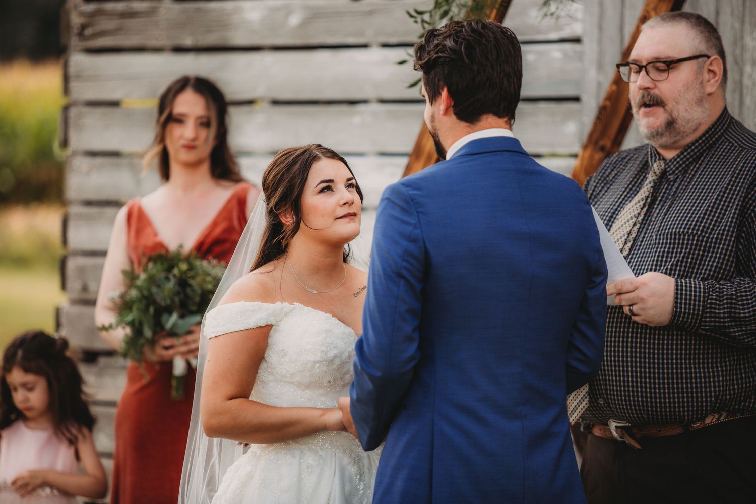 Couple stands before a rustic barn backdrop during their wedding ceremony with guests watching.