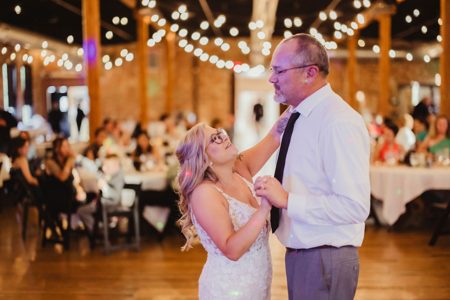 Guests dance together under twinkling lights in a rustic barn venue during a wedding celebration.