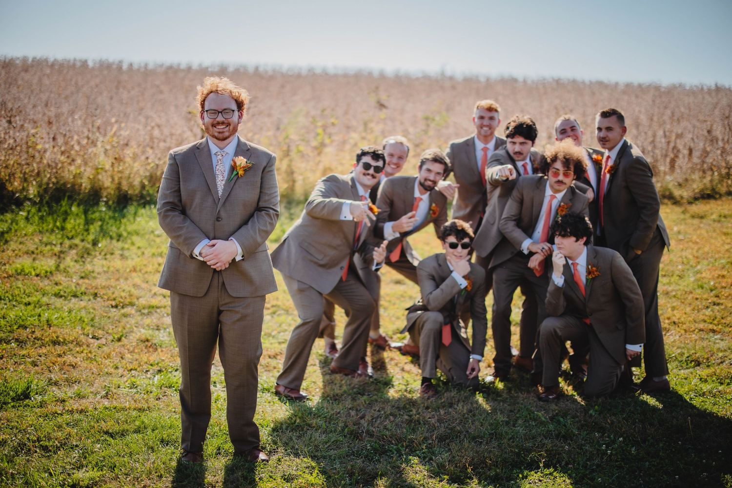 A lively group in formal attire poses playfully outdoors in a field with tall grass and autumn colors.