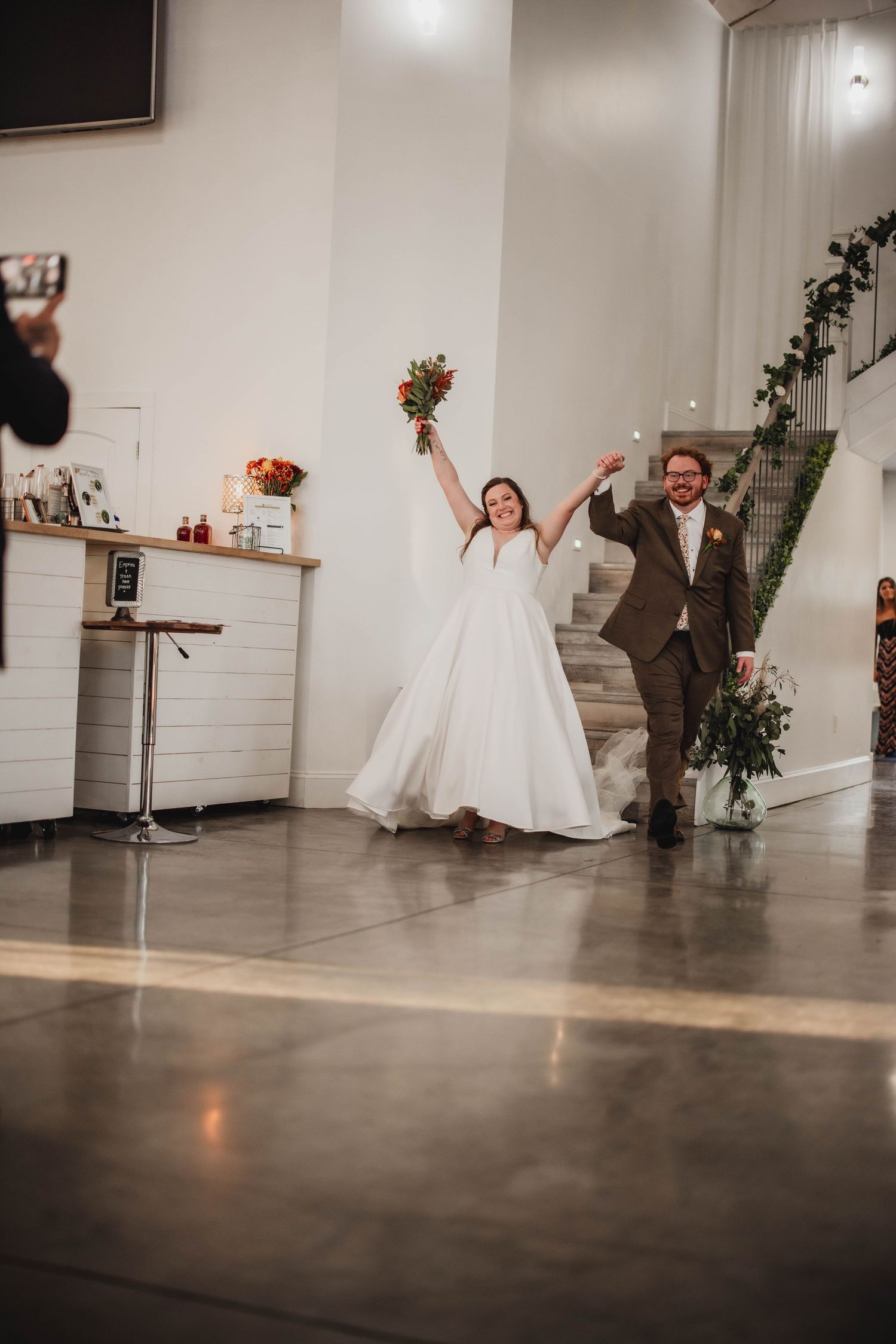 Couple shares an energetic entrance moment with raised arms and orange bouquets in a modern wedding venue.