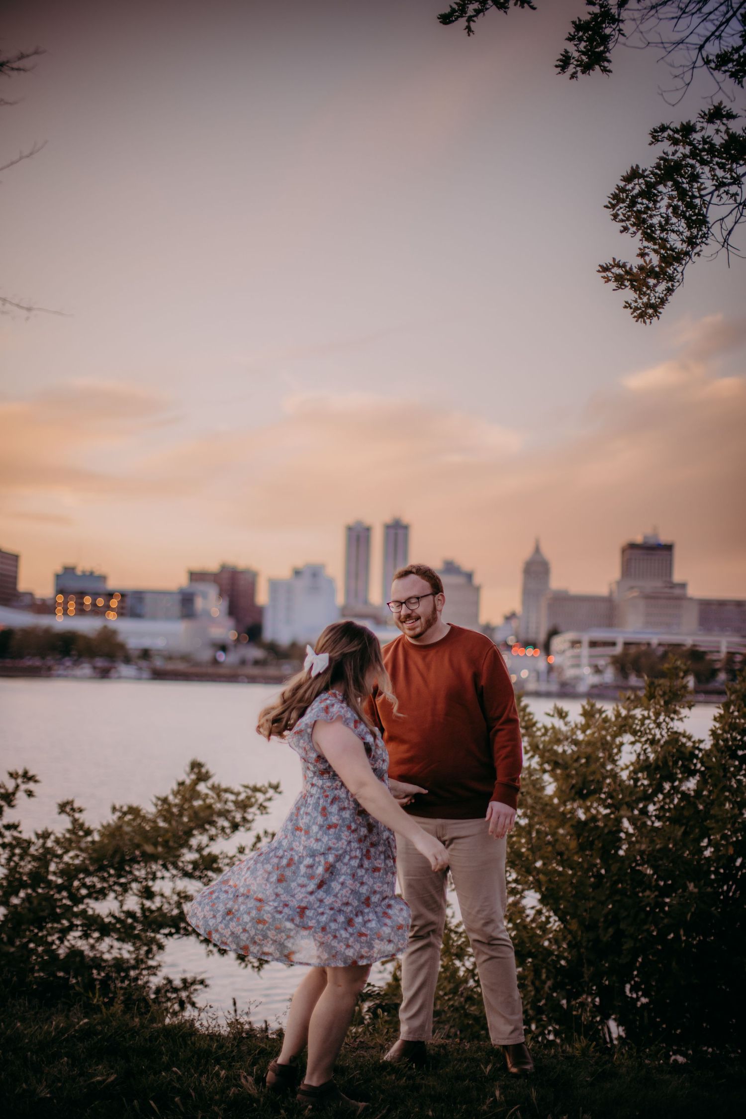 Couple shares playful moment on hill with waterfront cityscape during sunset.