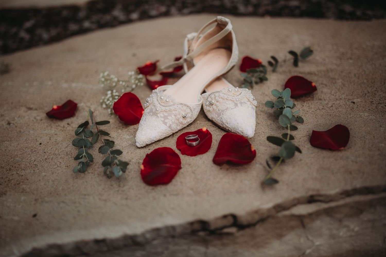Delicate white bridal shoes decorated with scattered rose petals and greenery on textured stone.