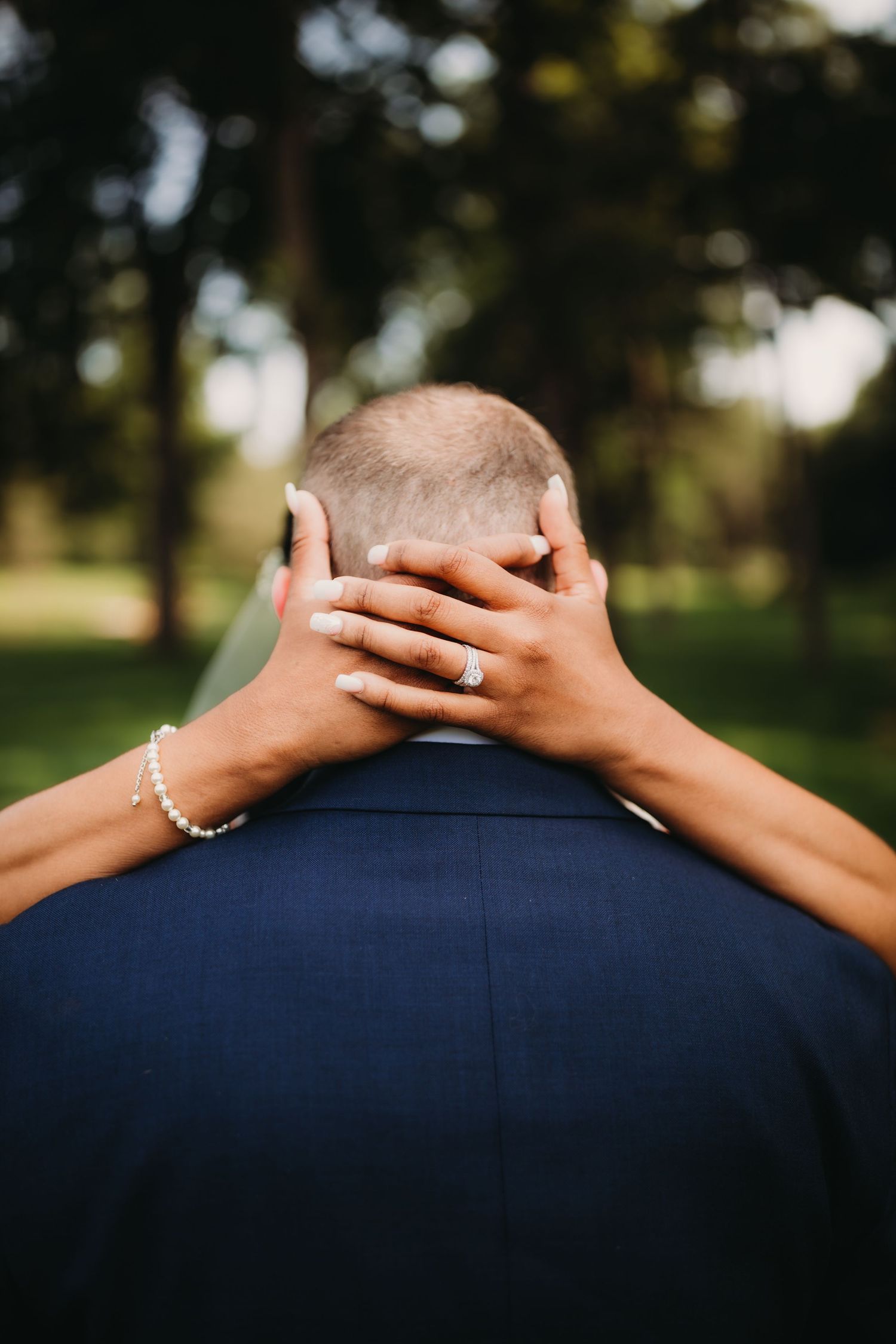 Close-up wedding photo showing bride's hands with rings embracing groom from behind in outdoor setting.