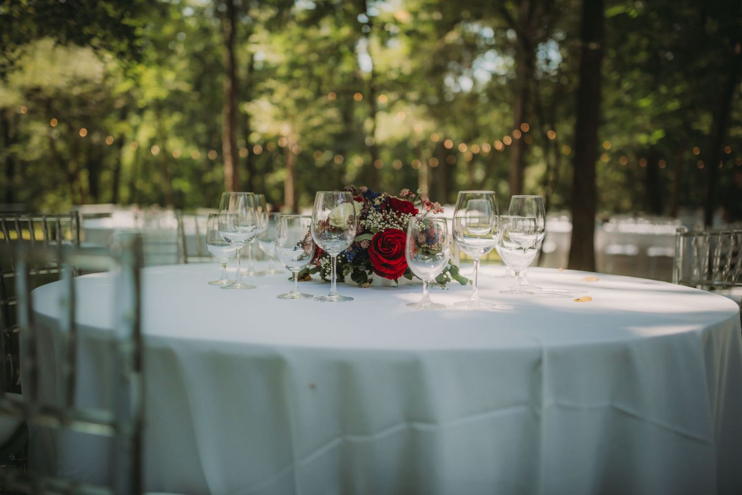 Elegant outdoor reception tables with white linens set beneath towering trees.