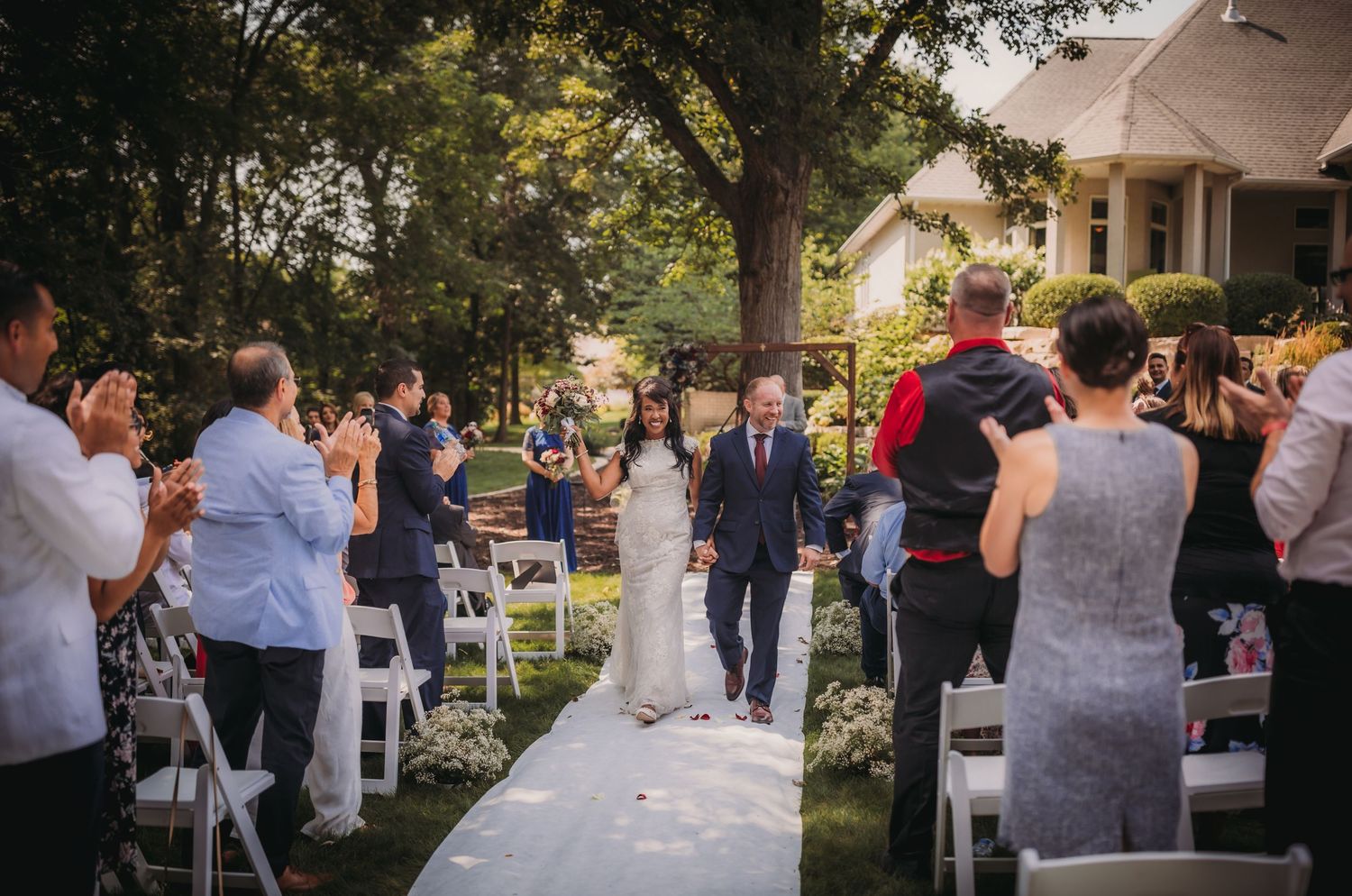 A garden wedding ceremony setup with guests mingling among white chairs and tall trees.