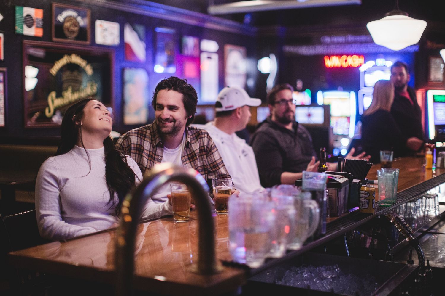 View down a bar counter showing customers enjoying drinks under neon lights.