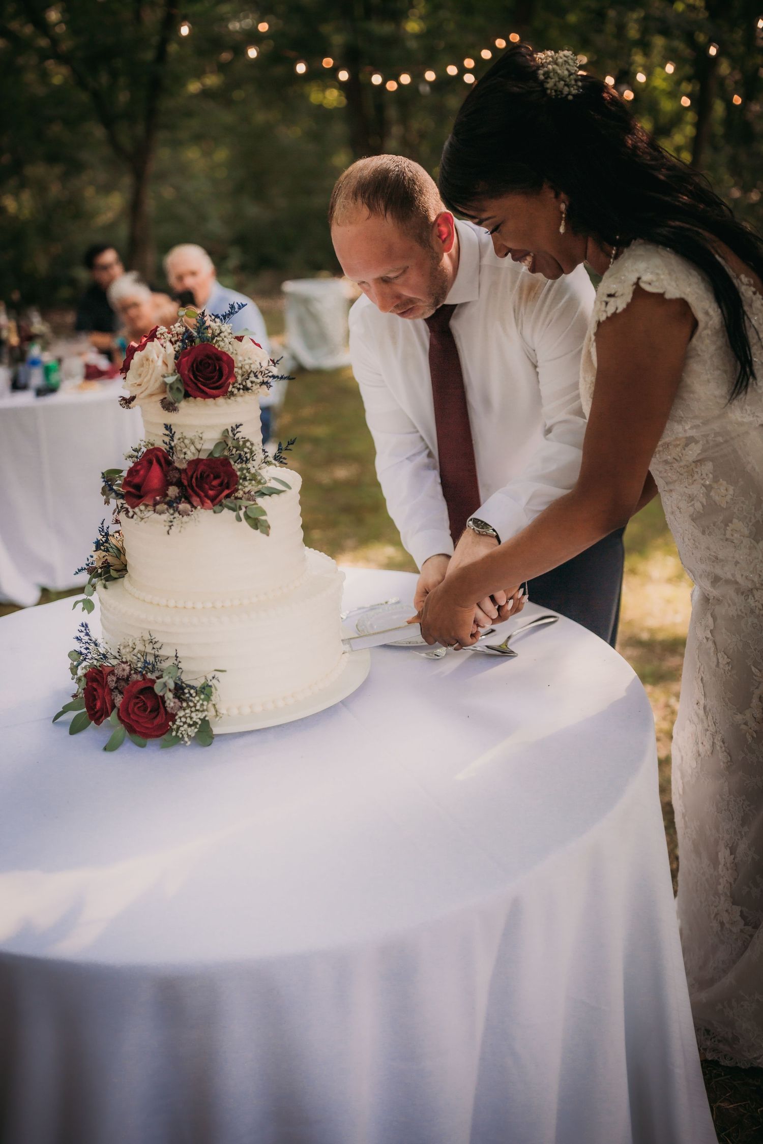 Wedding couple signing documents at an outdoor reception table with a white tiered cake decorated with red flowers.