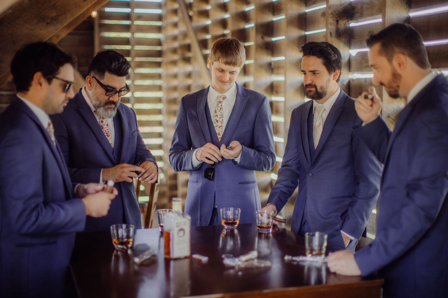 Groomsmen in navy suits gather around a table with drinks during pre-wedding preparations.