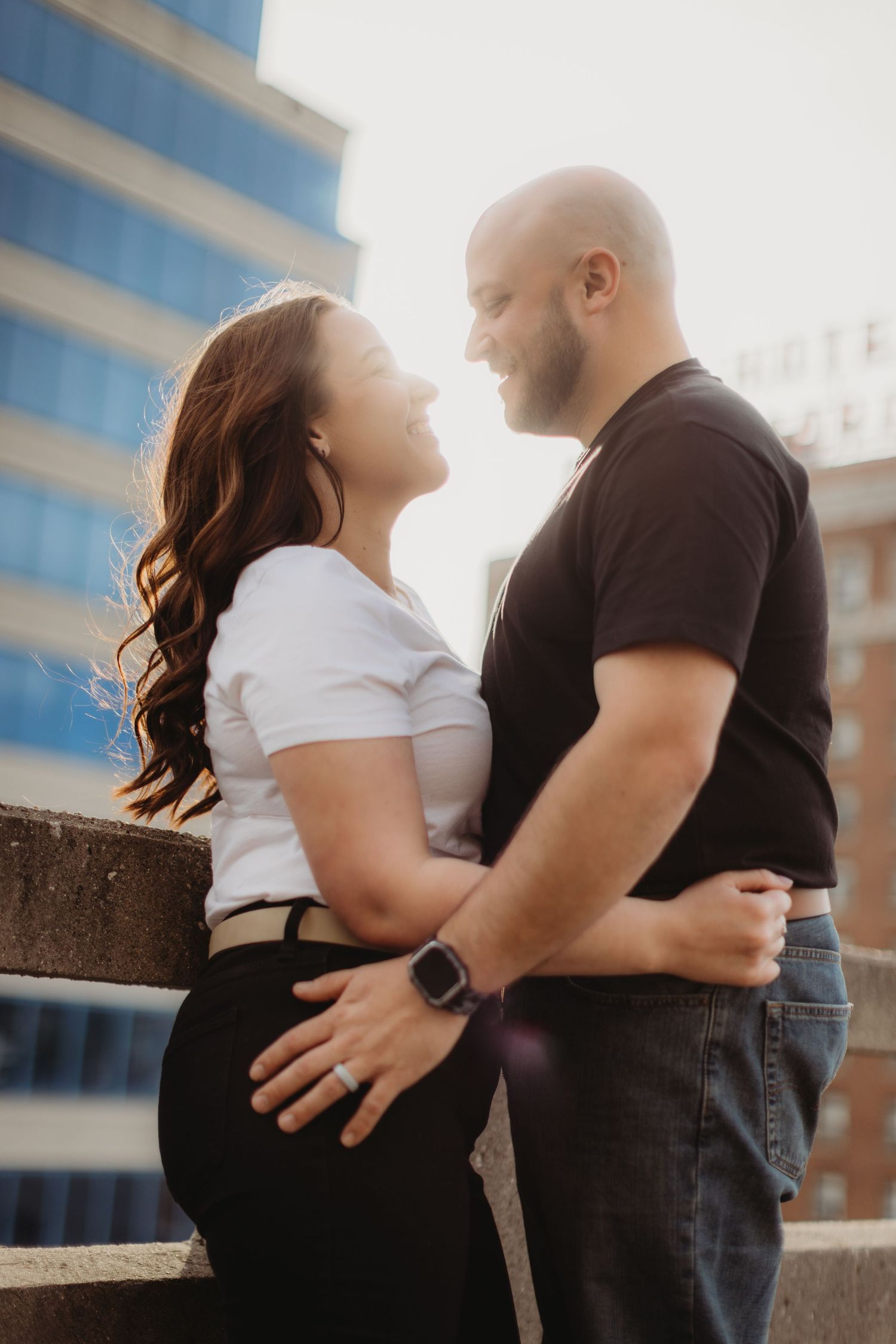 A couple shares an intimate moment against a concrete wall with skyline background.