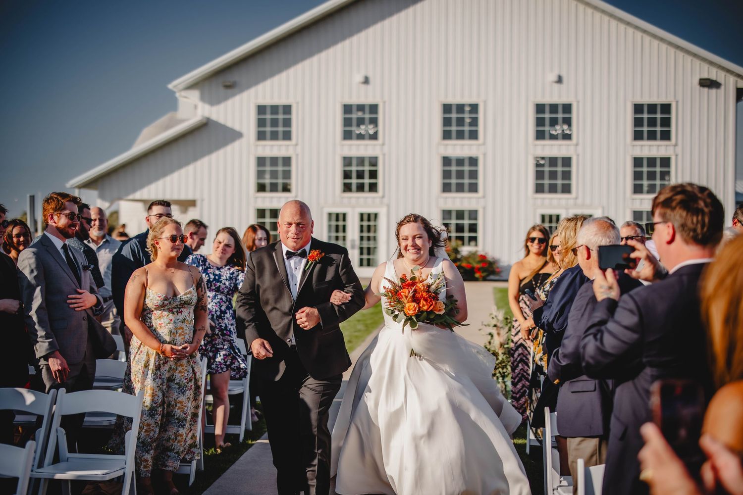 Bride walks down outdoor aisle with orange bouquet at rustic white barn wedding.