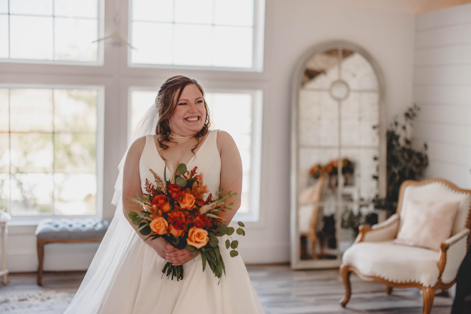 A bride clutches a stunning autumn-colored bouquet in a sun-filled bridal suite with vintage furniture.
