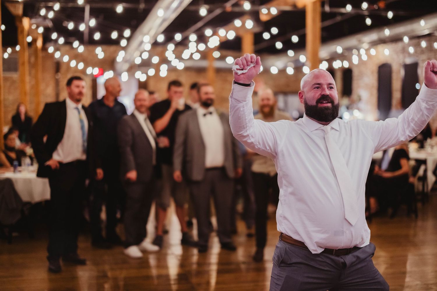 Celebratory dance moves on display at a rustic wedding reception under glowing string lights.
