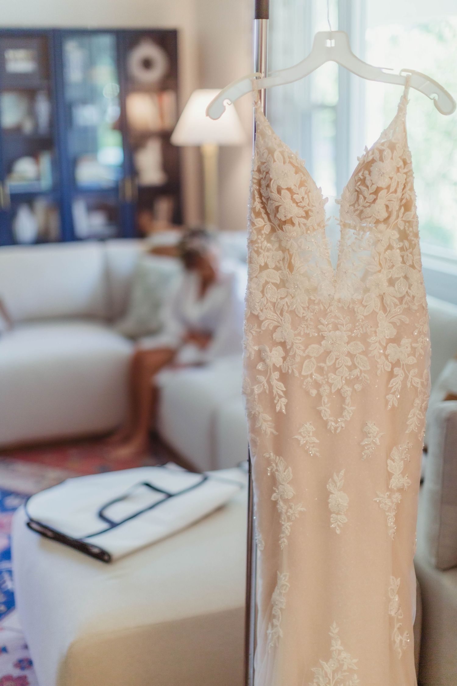Elegant cream lace wedding dress hanging by a window with blurred background.