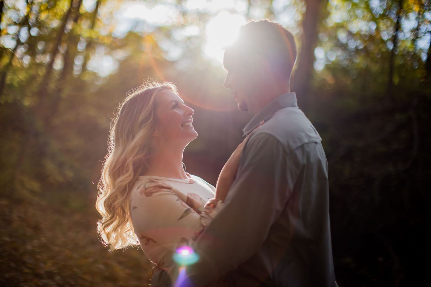 Couple in romantic embrace with dramatic sunlight streaming through trees creating lens flares.
