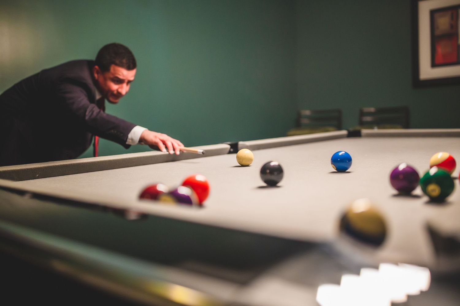 Person in dark clothing lines up a shot on a pool table with colorful billiard balls scattered across the felt.