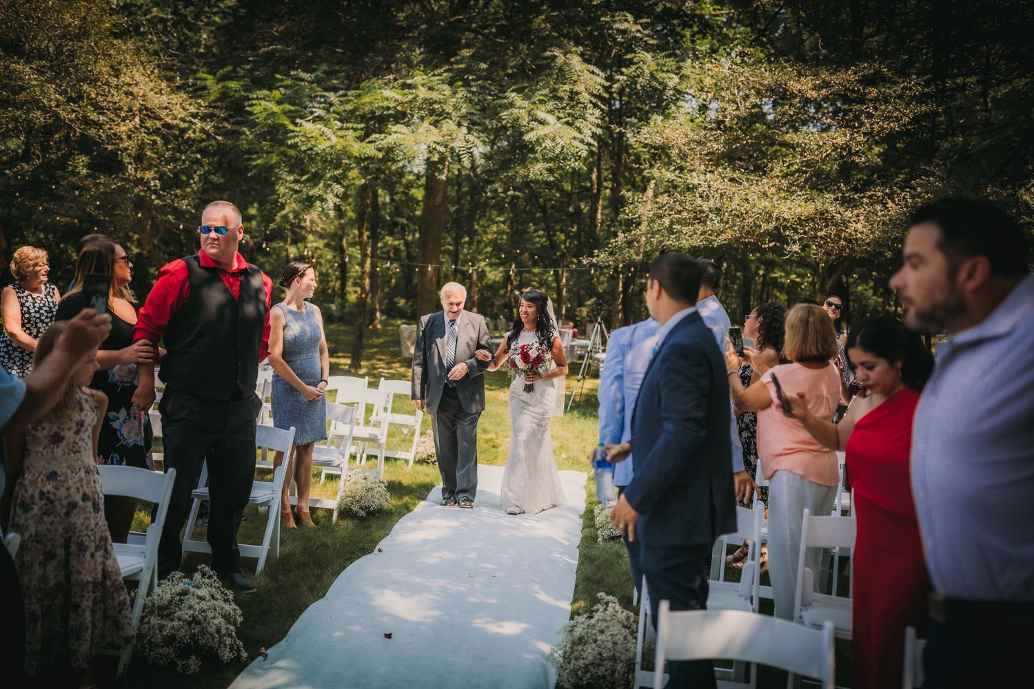 Outdoor wedding ceremony setup in a wooded area with white chairs and floral decorations.