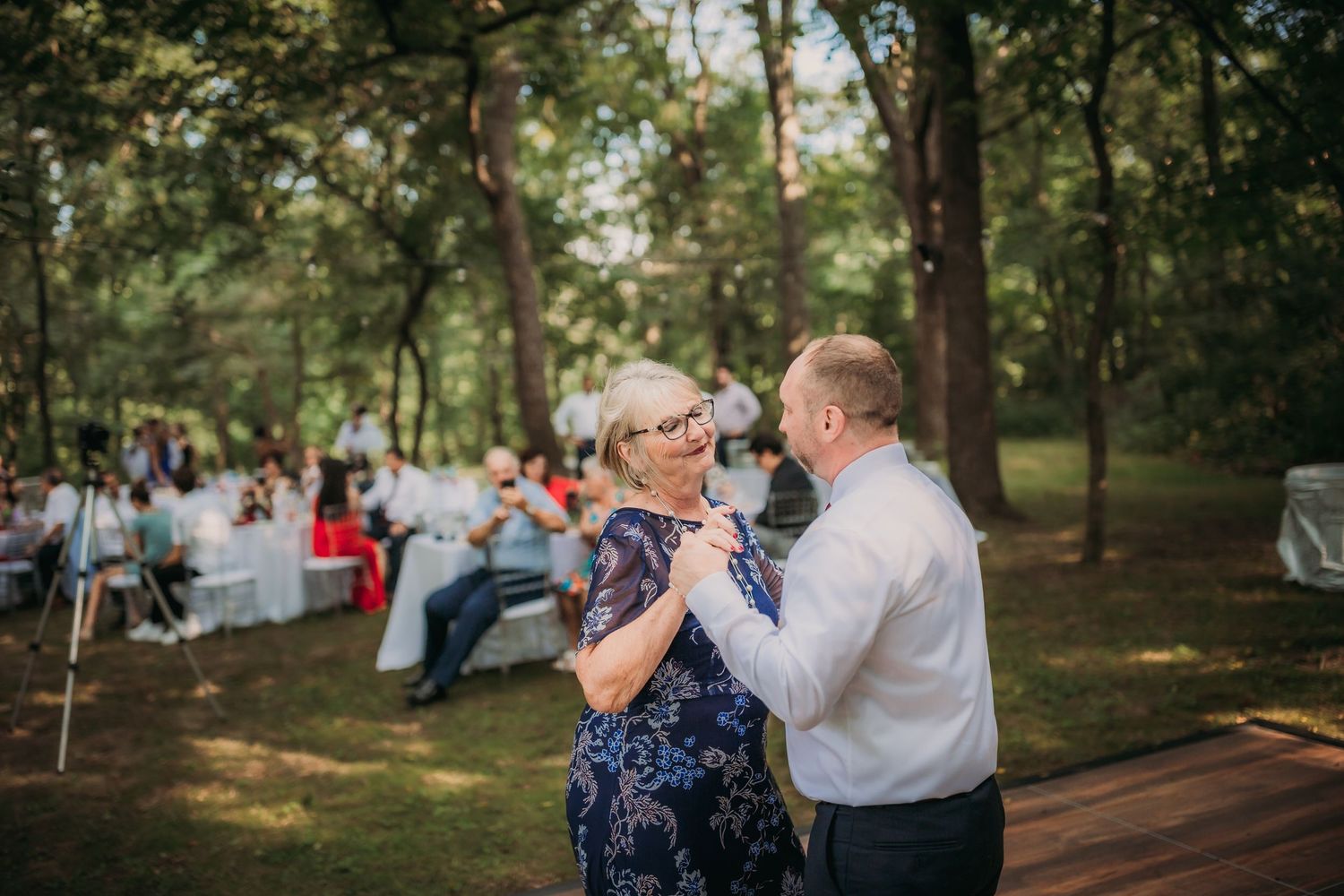Couples share a dance on a wooden platform at an outdoor woodland reception with string lights overhead.