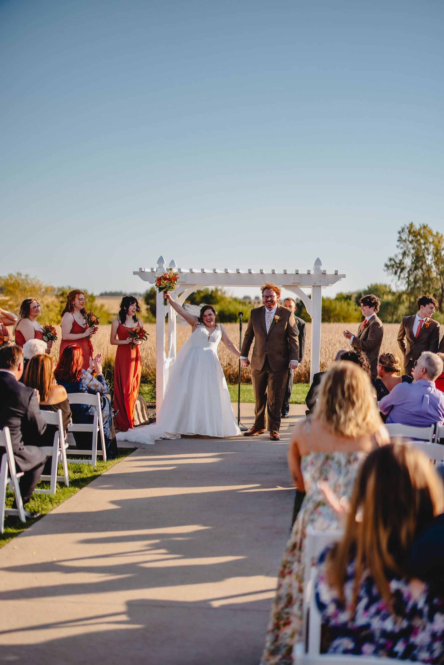 Newlyweds celebrate with raised hands under a white pergola during outdoor wedding ceremony.