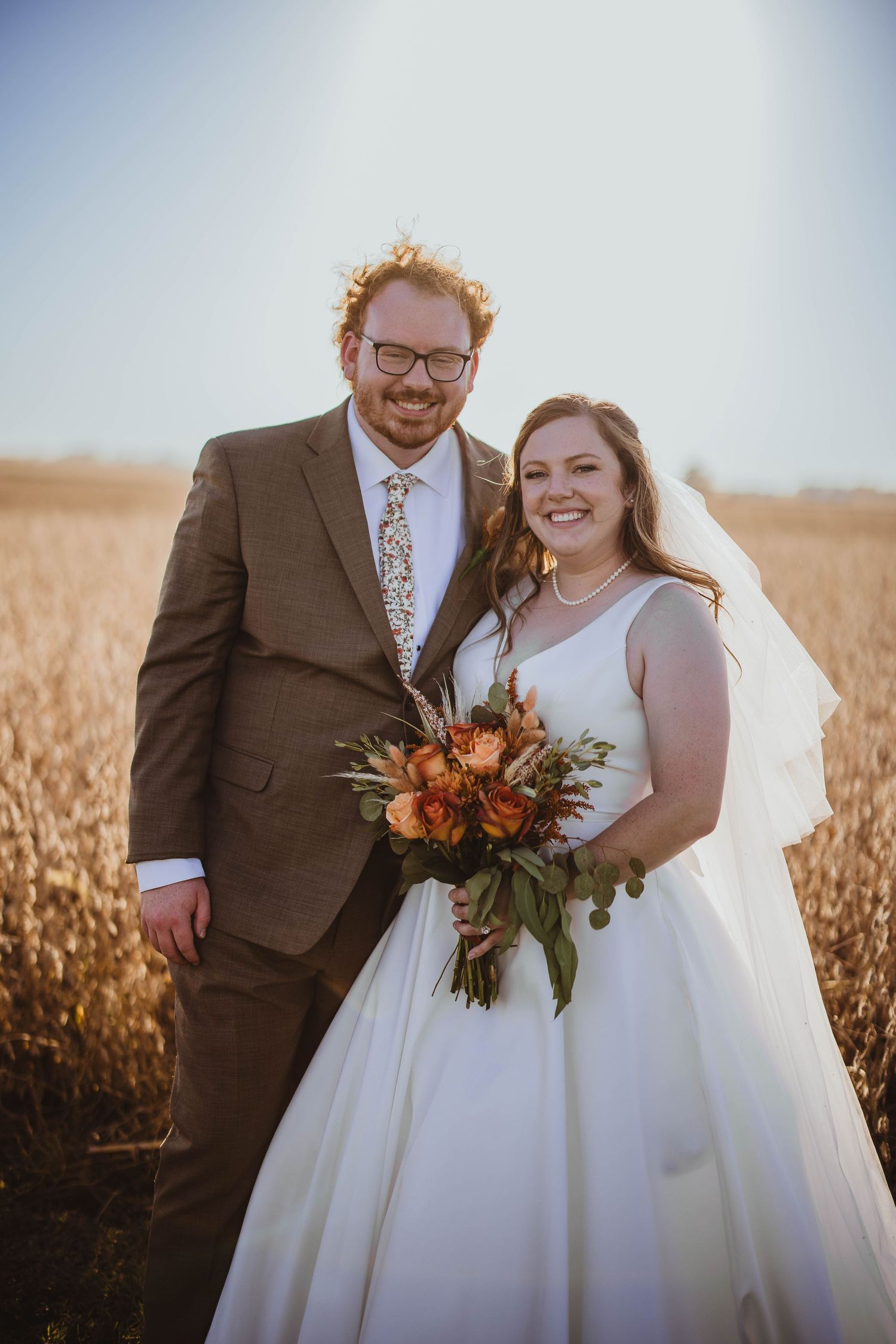 Newlyweds posing in a golden wheat field at sunset with autumn-colored wedding bouquet.