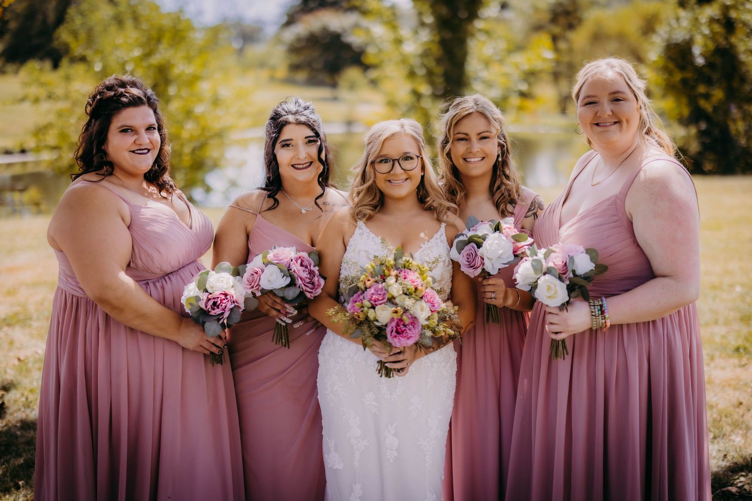 Wedding party poses with pink and white floral bouquets in rose-colored dresses.