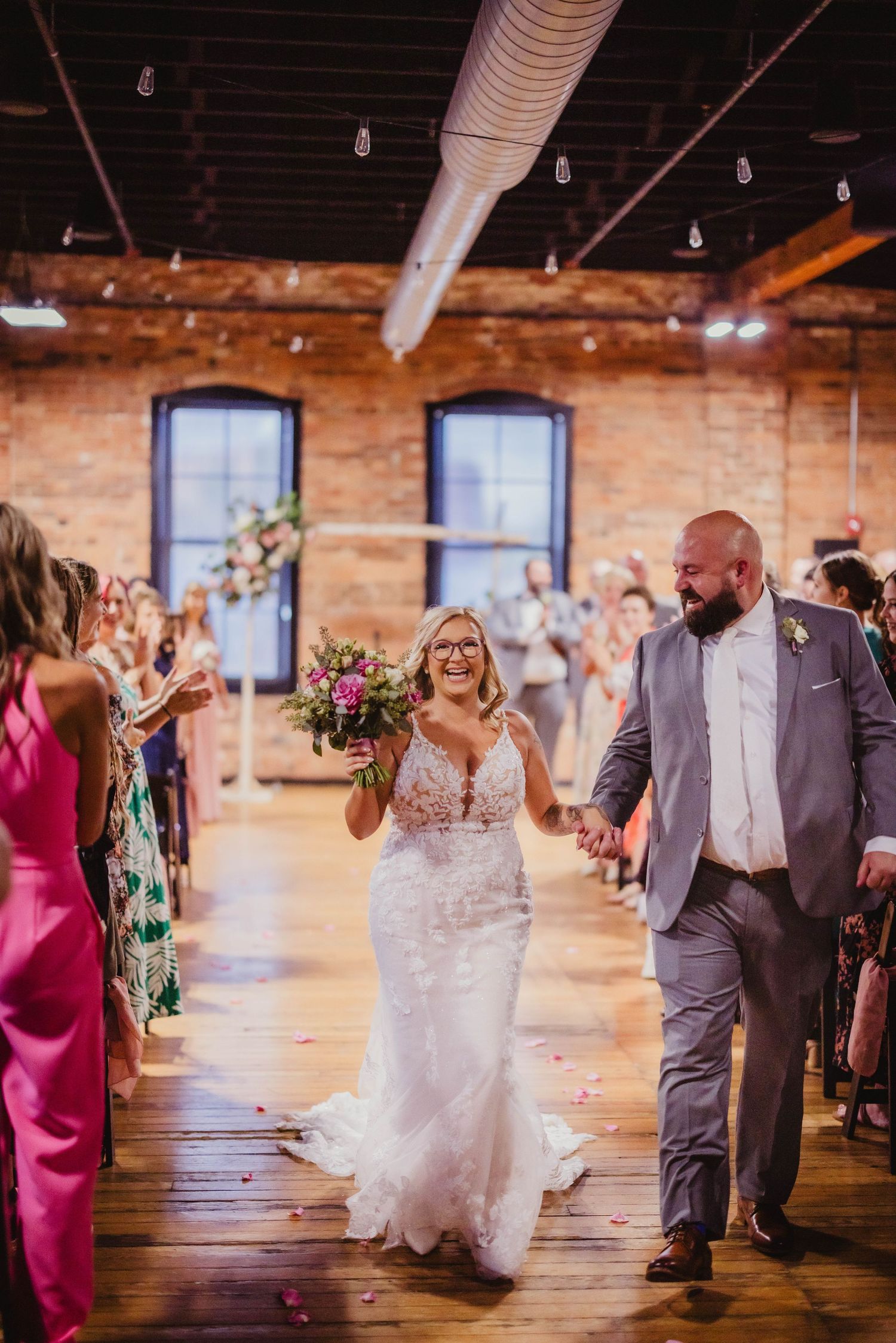 Joyful wedding recessional in brick-walled venue with guests cheering and flowers decorating windows.