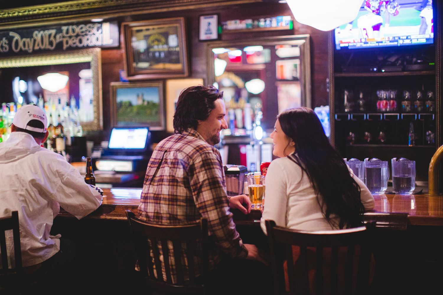 Couple enjoying conversation at a dimly lit bar counter with neon signs and televisions in the background.
