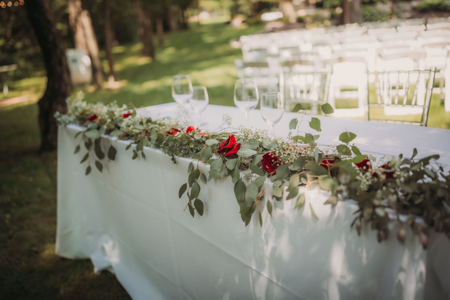 Elegant wedding table setup with a white tablecloth decorated with greenery and red roses in an outdoor garden setting.