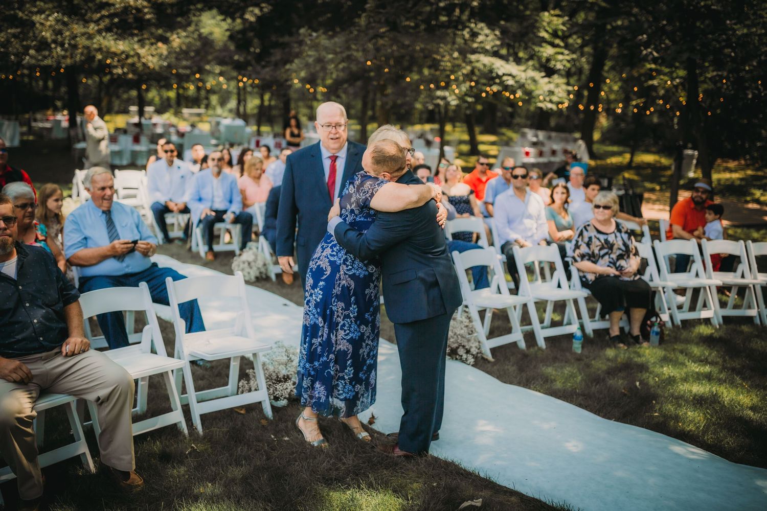 Guests watch an intimate moment at an outdoor wedding with white folding chairs and trees.