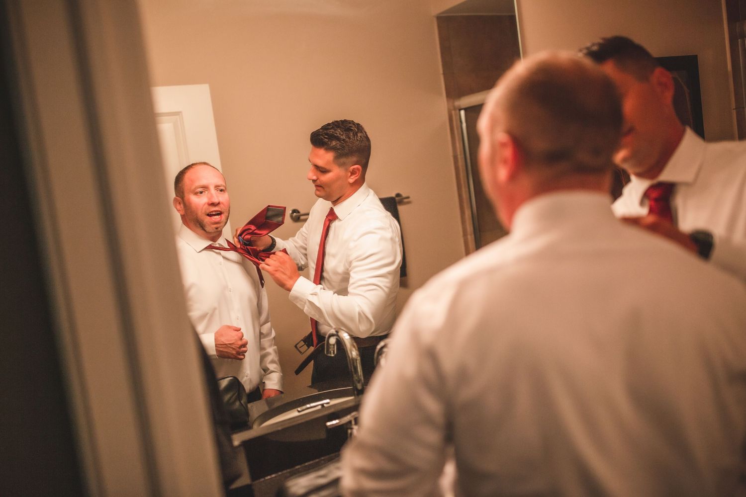 Group of men in formal attire sharing a laugh while getting ready in a bathroom mirror.