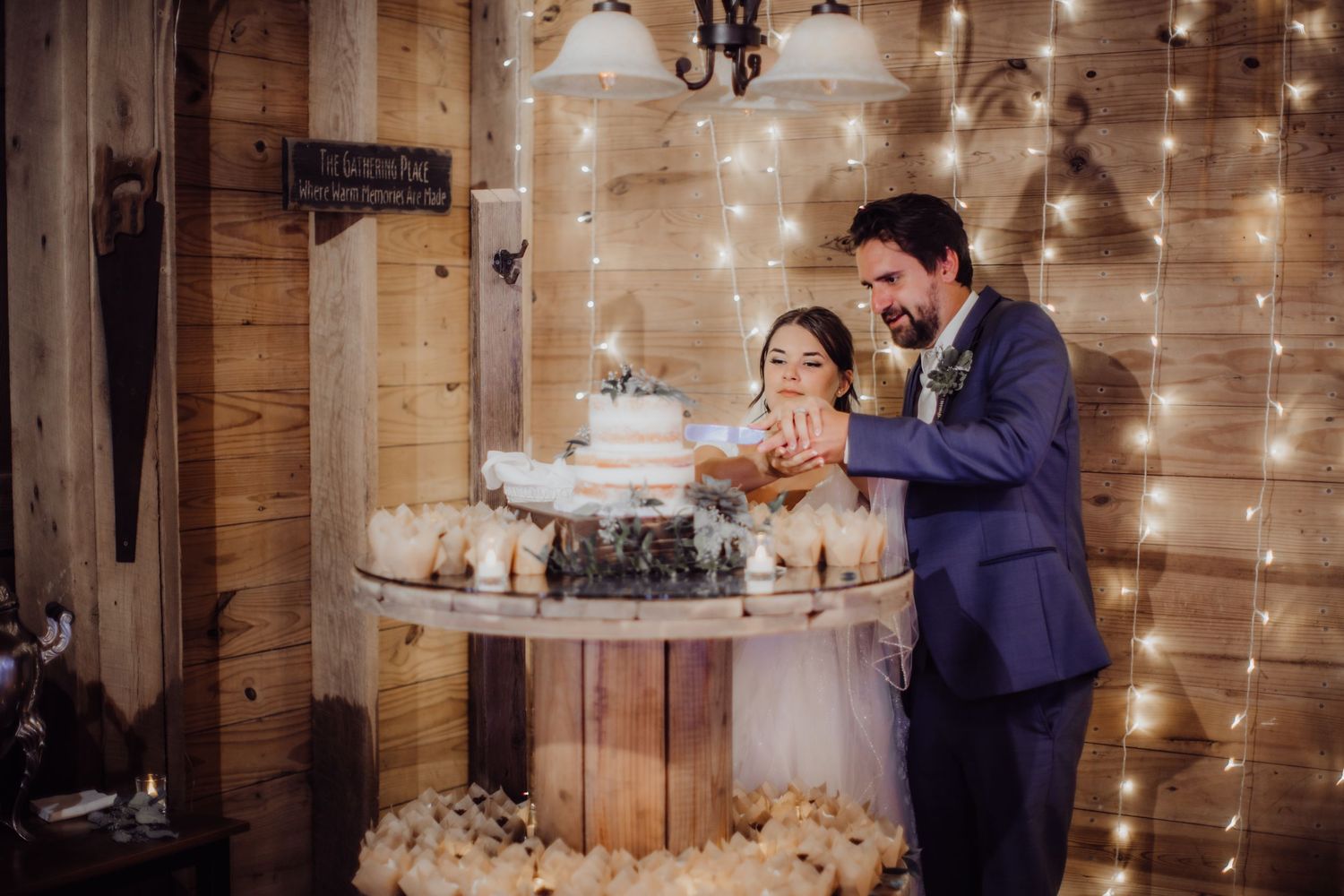 A joyful cake cutting moment at a rustic barn wedding reception with twinkling lights in the background.