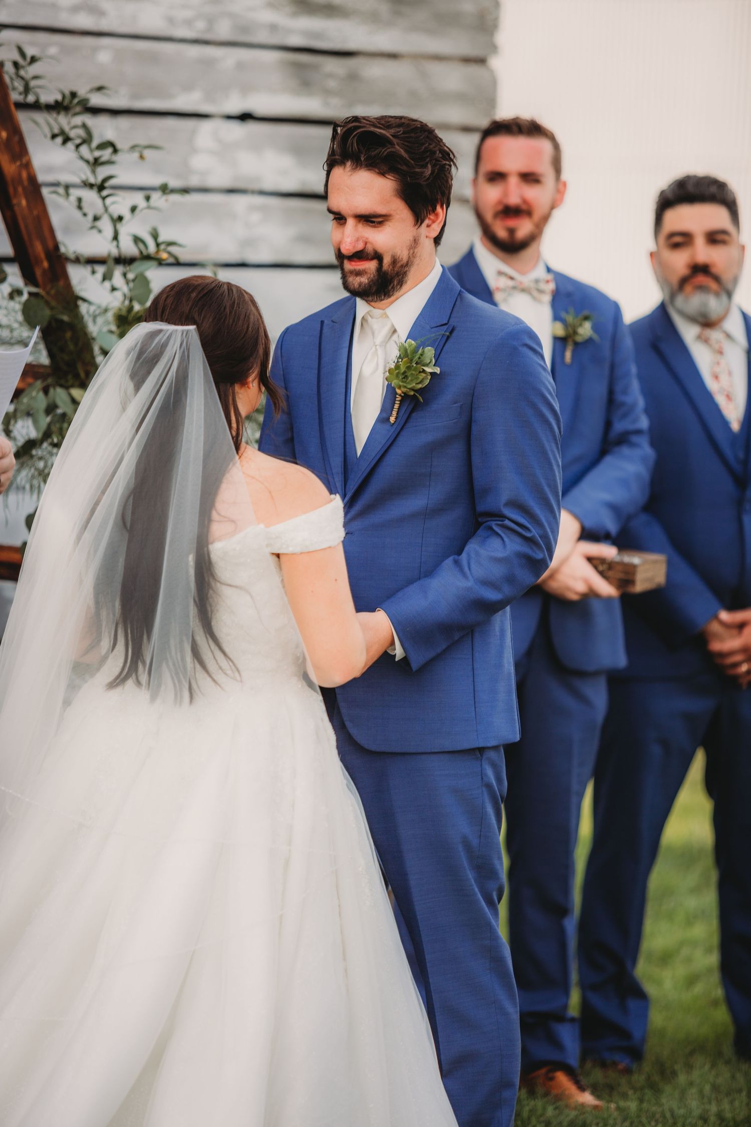Groomsmen in matching navy suits watch the wedding ceremony from beside a rustic barn.
