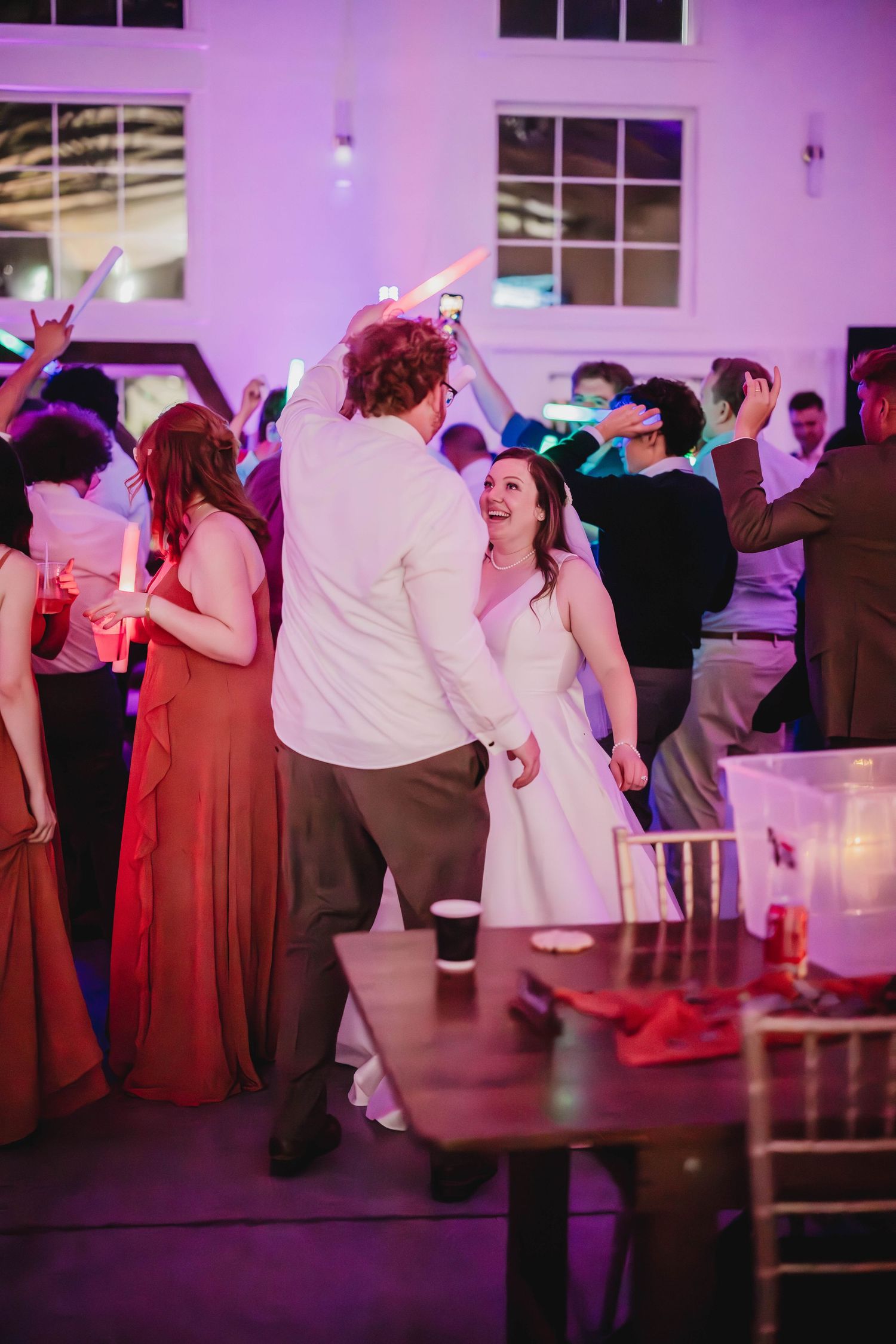 Wedding guests dance at a reception under purple lighting while a couple enjoys their first dance.