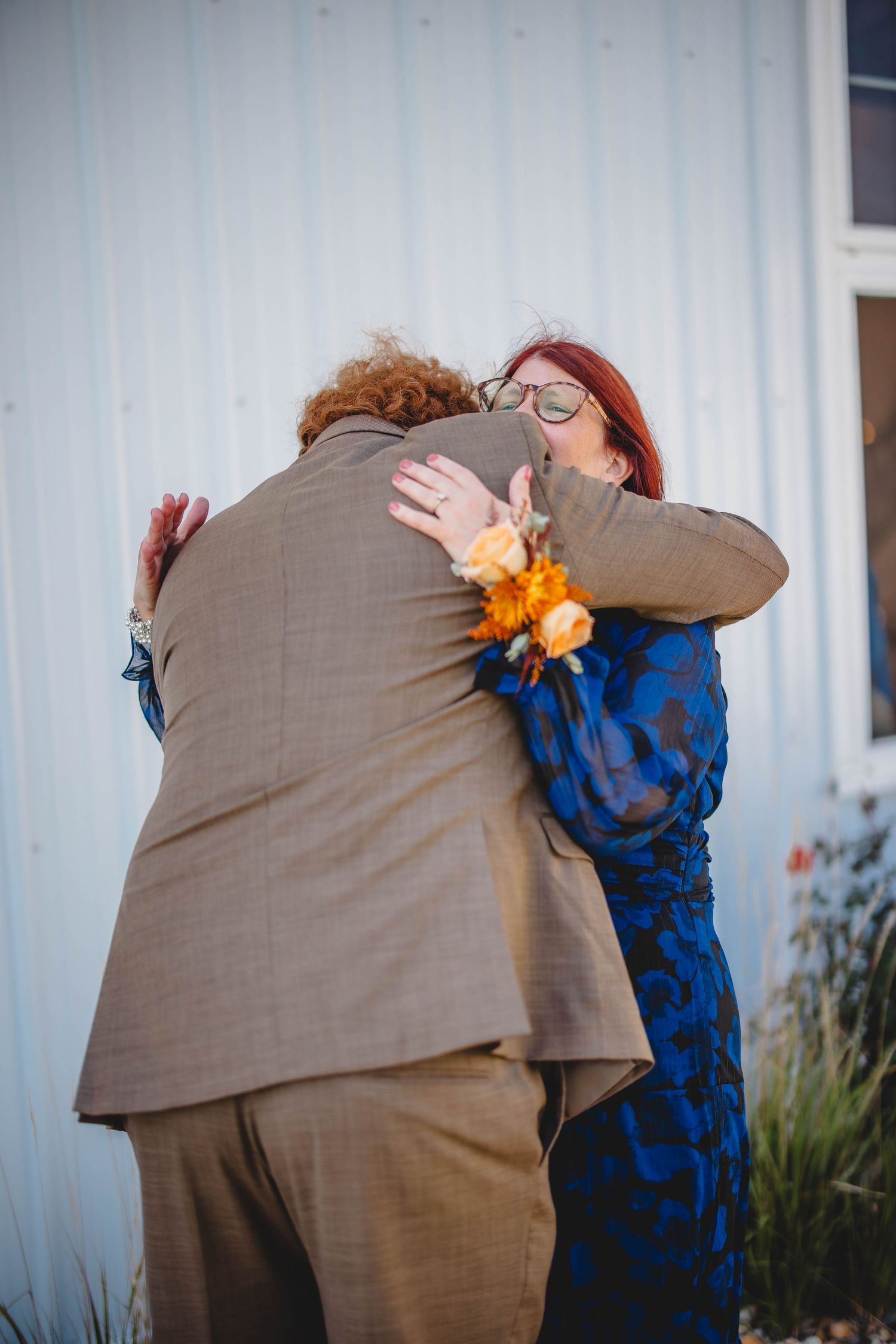 A couple sharing warm embrace outside white building with orange boutonniere visible.