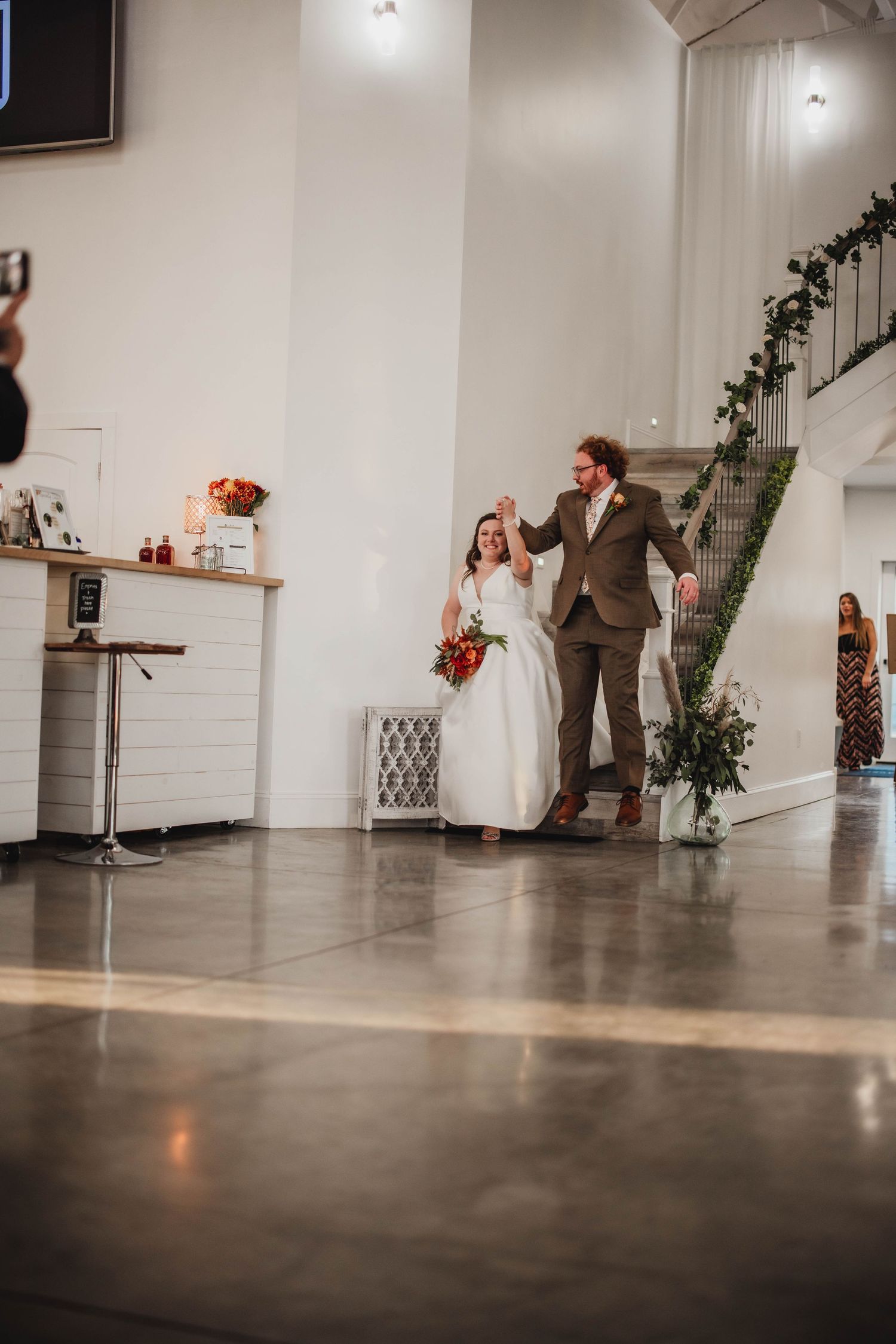 Newlyweds make their grand entrance into an elegant white venue with polished concrete floors and greenery-adorned stairs.