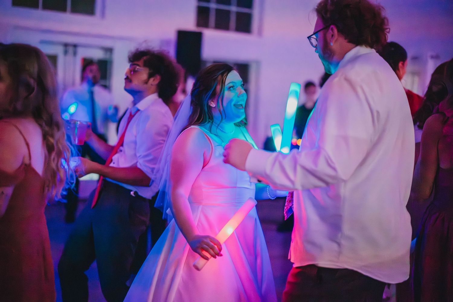 Partygoers dancing under colorful pink and blue lighting at an indoor reception.