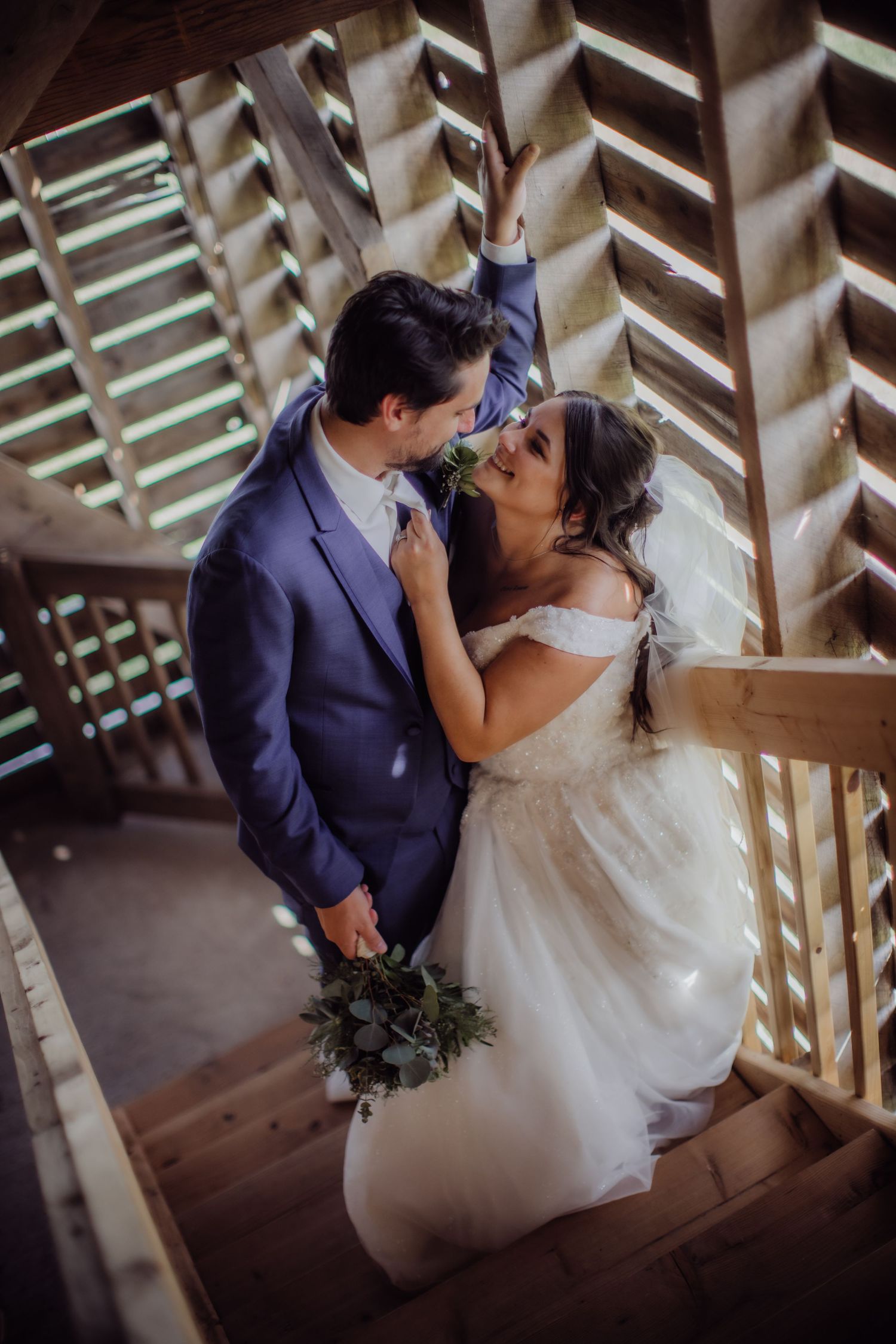 Bride in white gown and groom in navy suit embrace on rustic wooden staircase.