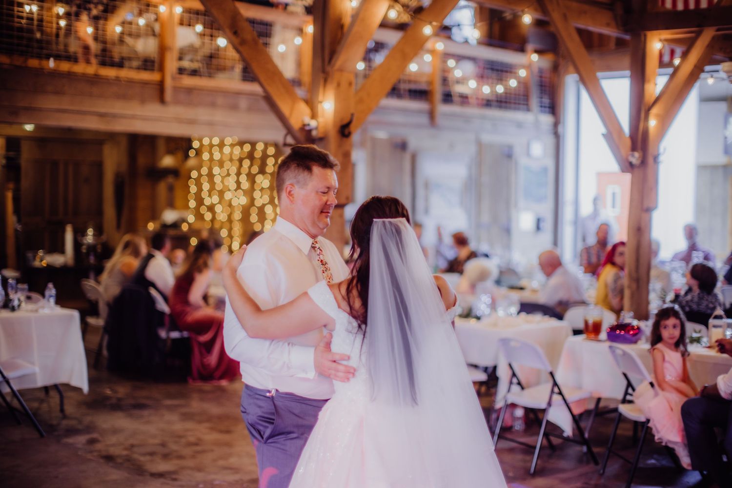 Couple shares an intimate dance moment in a rustic reception hall with string lights.