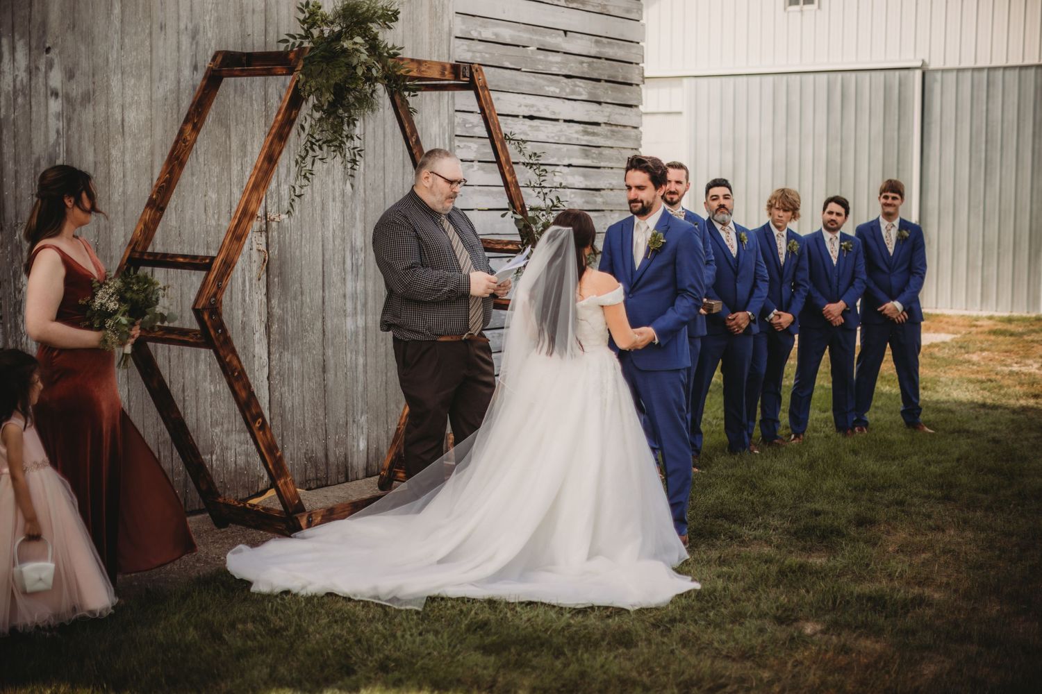 Wedding party posed in front of rustic barn with wooden hexagonal arch.