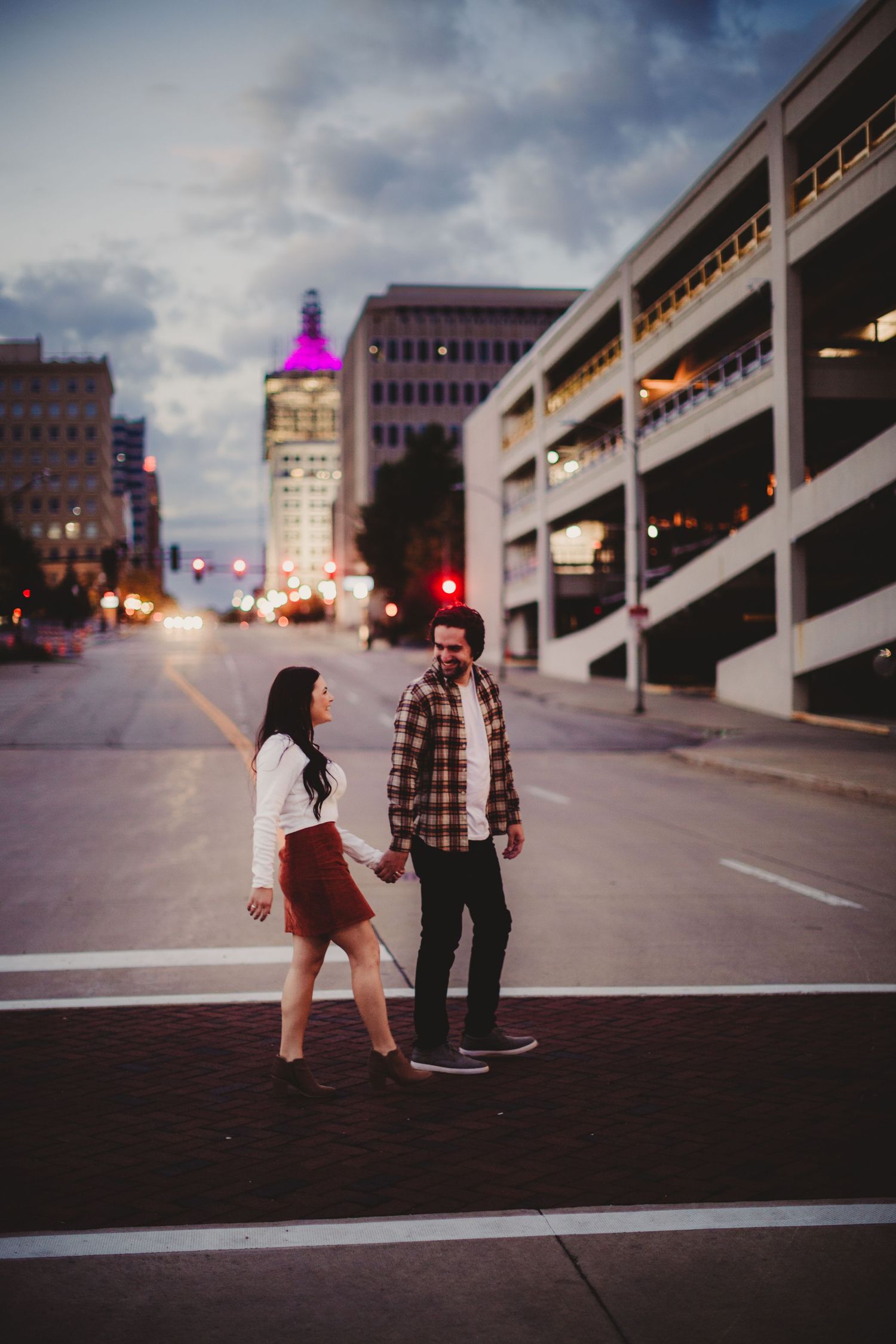 A couple stands in front of a parking structure at dusk with a colorfully lit tower in the background.