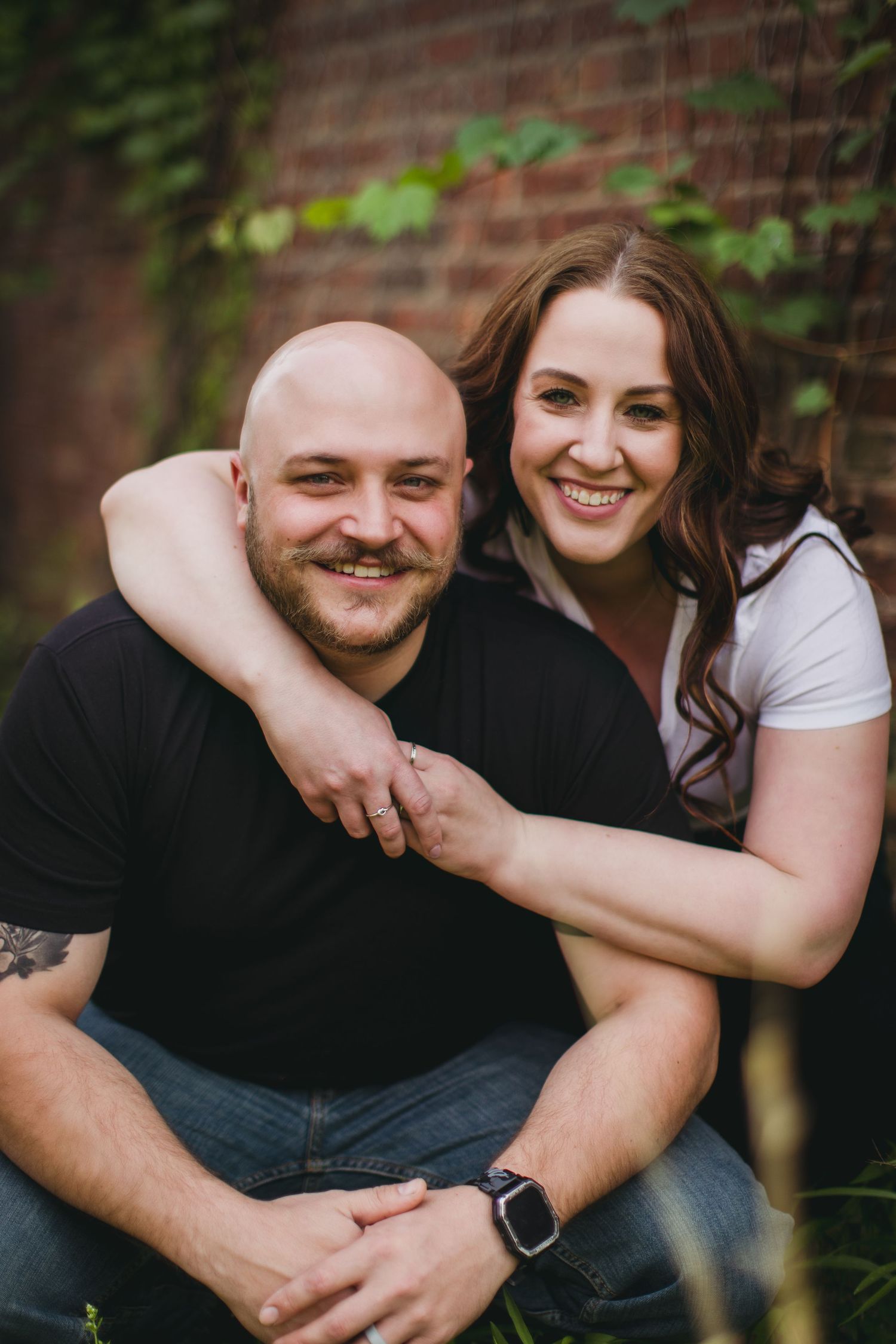 Candid engagement photos showing couple laughing and embracing near brick wall with greenery.