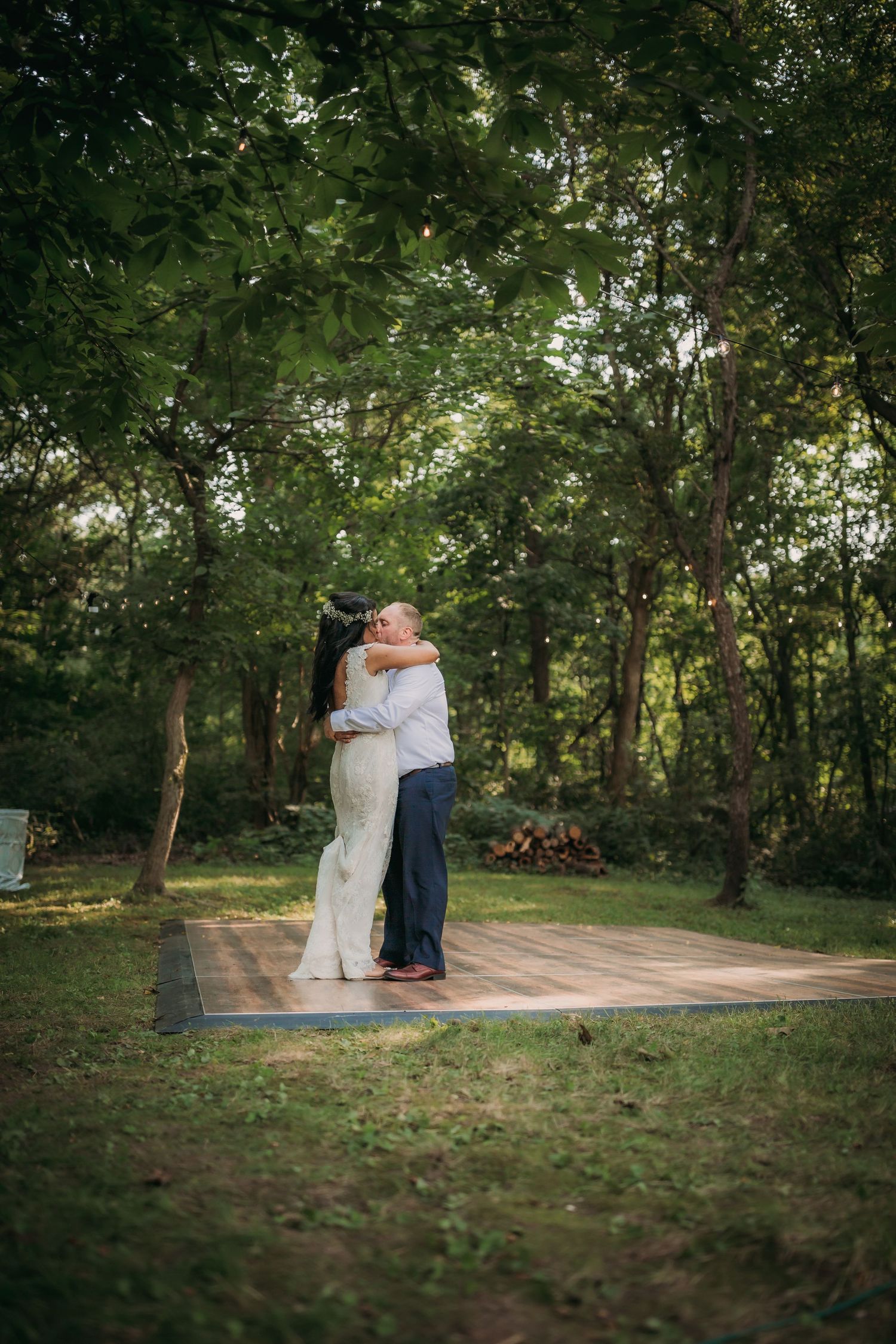 Couple sharing a romantic embrace on a wooden platform surrounded by lush green trees in an outdoor setting.