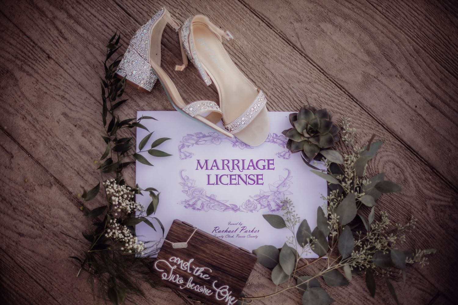 Marriage license document with wedding shoes and eucalyptus on wooden floor.